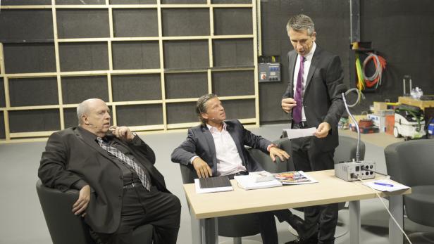 Drei Männer in Anzügen sitzen oder stehen an einem Tisch in einem Studio.