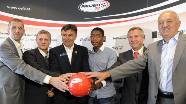 Mehrere Personen in Anzügen halten einen roten Fußball mit der Aufschrift „Projekt 12“.