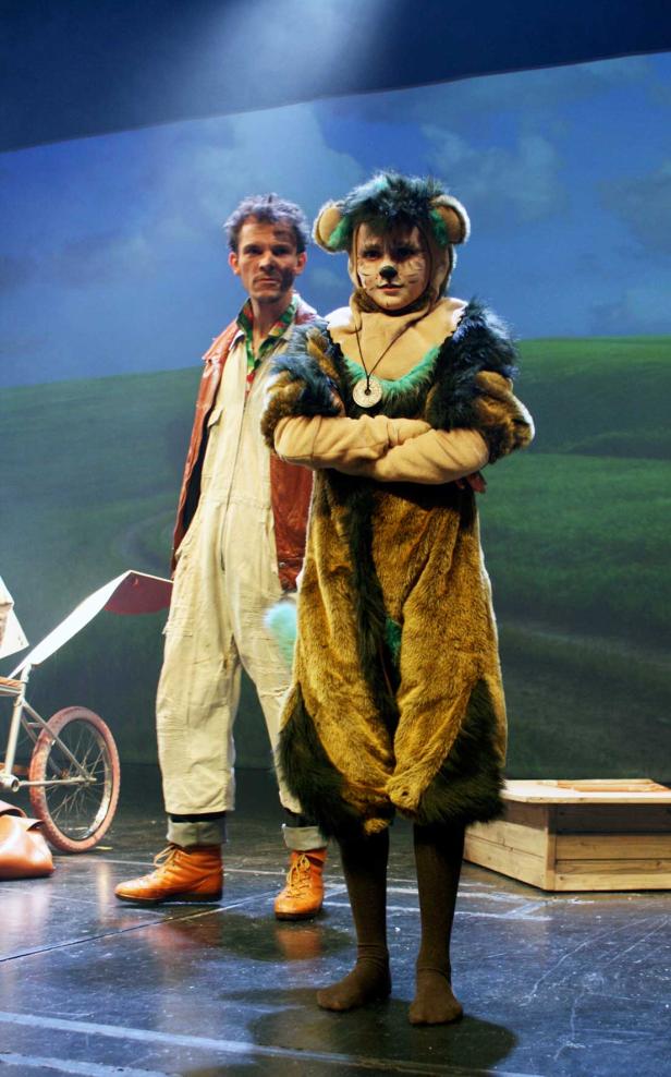 Ein Schauspieler im Löwenkostüm steht mit verschränkten Armen auf einer Bühne, neben ihm ein Mann in Arbeitskleidung.