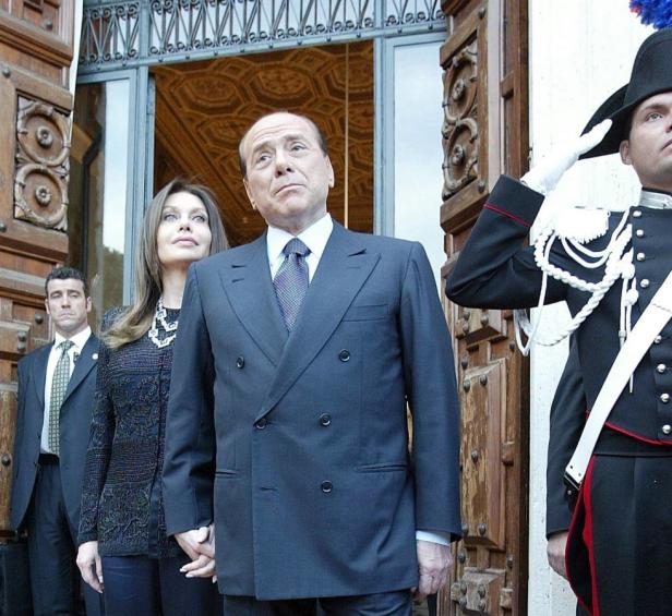Silvio Berlusconi mit einer Frau vor einem Gebäude, flankiert von Sicherheitsleuten und einem Soldaten.