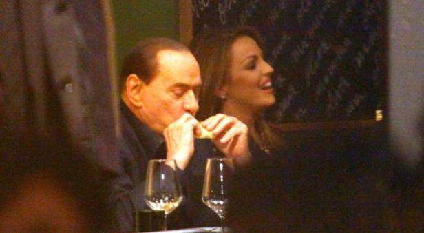 Silvio Berlusconi isst in einem Restaurant, während eine Frau lächelt.