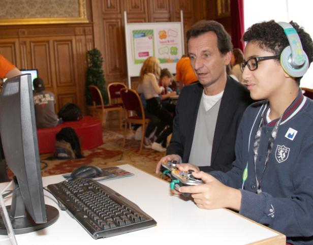 Ein Jugendlicher mit Kopfhörern spielt mit einem Controller vor einem Monitor, während ein Mann zusieht.
