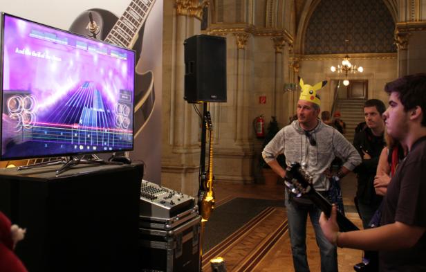 Ein Mann mit Pikachu-Hut steht vor einem Bildschirm, der ein Gitarrenspiel zeigt.
