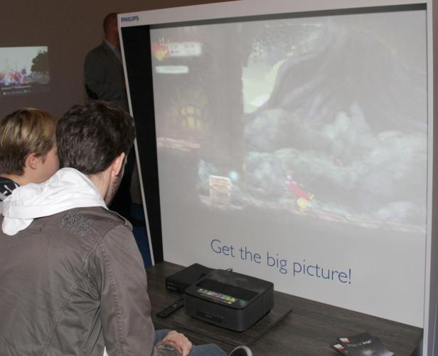 Zwei Personen sehen einem Mann beim Spielen eines Videospiels auf einem Philips-Projektor zu.