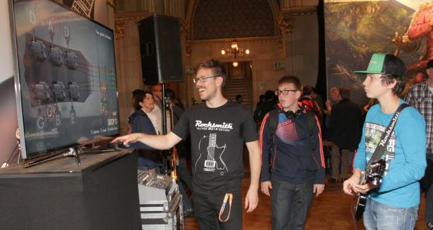 Ein Mann erklärt das Spiel „Rocksmith“ auf einem Bildschirm, während Jugendliche zusehen.
