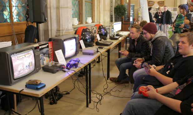 Mehrere Personen spielen an alten Konsolen und Röhrenfernsehern Videospiele.