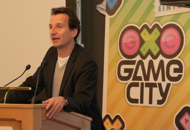 Ein Mann spricht auf einer Bühne vor einem „OXO Game City“-Banner.