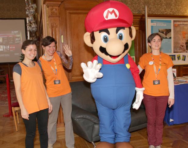 Ein Mario-Maskottchen posiert mit drei Frauen in orangefarbenen T-Shirts.