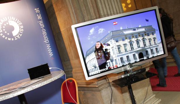 Ein Bildschirm zeigt eine Person vor einem Gebäude mit der österreichischen und der EU-Flagge.