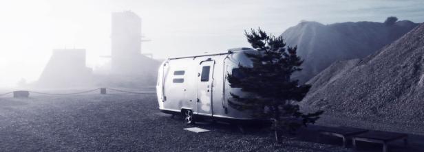 Ein silberner Wohnwagen steht vor einer nebligen Industrielandschaft.