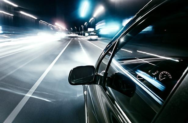 Ein Auto fährt nachts mit hoher Geschwindigkeit auf einer beleuchteten Straße, Lichter ziehen Spuren.