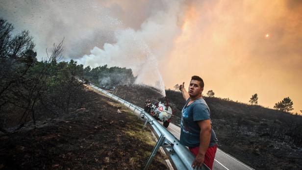 Ein Mann steht an einer Straße und zeigt auf ein Feuerwehrauto, das einen Waldbrand bekämpft.