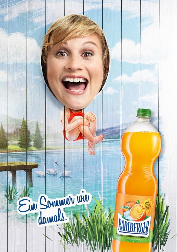 Radlberger - Kampagne - Sujet 4 - Ortner&Weihs Eine Frau schaut durch ein Loch in einer Holzwand, im Hintergrund ein See und eine Flasche Radlberger Orange.