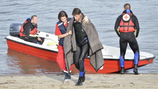 Ein Retter hilft einem durchnässten Mann mit einer Decke am Ufer, während andere Rettungskräfte im Hintergrund zu sehen sind.