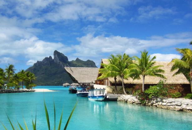 Bungalows mit Strohdächern und Boote auf dem türkisfarbenen Wasser vor einer Bergkulisse.