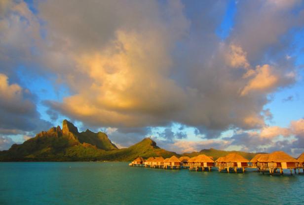 Bungalows auf dem Wasser vor einer tropischen Insel unter einem bewölkten Himmel.
