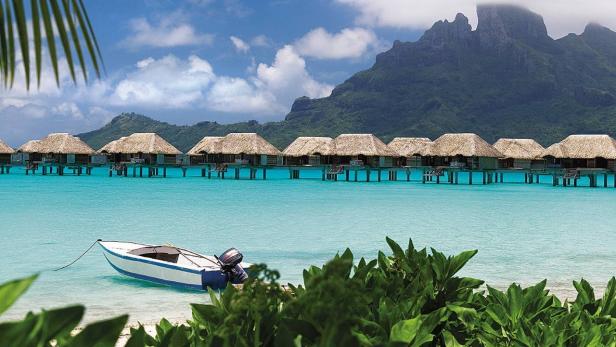 Bungalows mit Strohdächern stehen auf Stelzen im türkisfarbenen Wasser, im Vordergrund ein Boot.