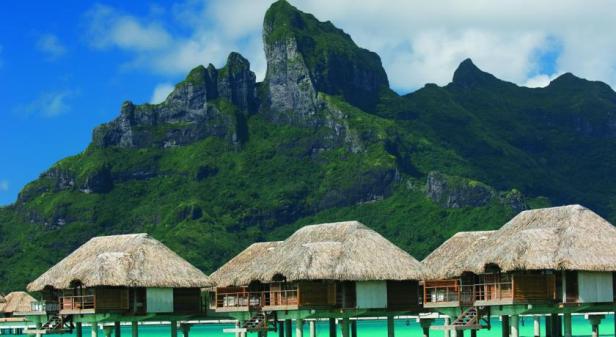Bungalows mit Strohdächern stehen auf Stelzen im türkisfarbenen Wasser vor einem grünen Berg.