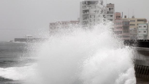 Eine hohe Welle bricht an einer Küstenmauer vor einer Stadt.