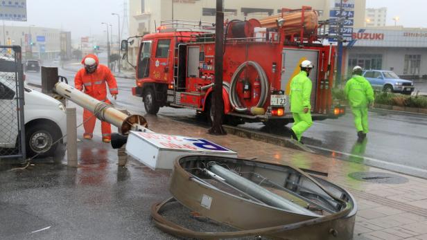 Ein umgestürztes Schild liegt auf der Straße, während Einsatzkräfte in Regenkleidung und ein Feuerwehrauto zu sehen sind.