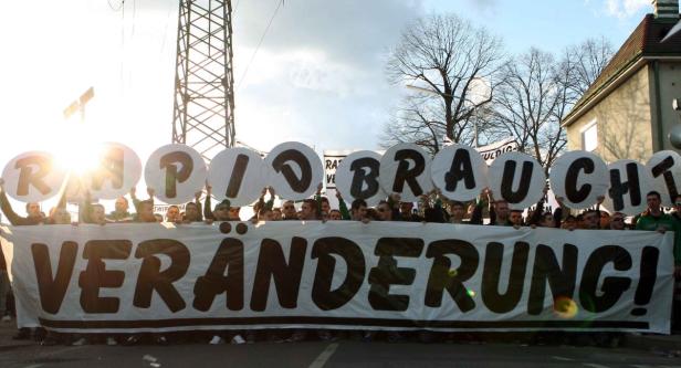 Eine Gruppe von Menschen demonstriert mit einem Banner, auf dem „Veränderung“ steht, und Schildern mit der Aufschrift „Rapid braucht“.