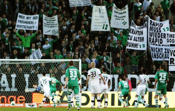 Fußballspiel mit Spielern in Grün und Weiß vor einer Zuschauermenge mit Transparenten.