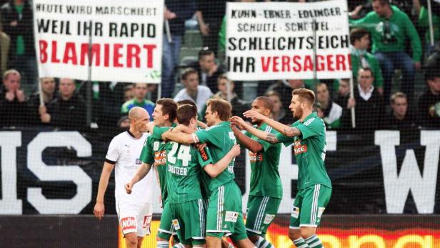 Fußballspieler in Grün feiern vor dem Hintergrund von Spruchbändern, die Rapid Wien verspotten.