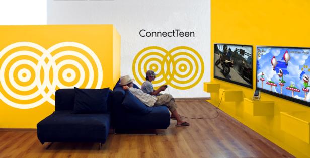 Zwei Jugendliche sitzen auf einem Sofa und spielen Videospiele vor zwei Bildschirmen.