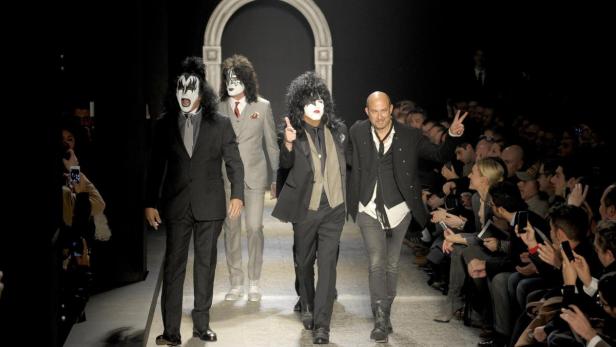 Männer in Anzügen und mit KISS-Make-up laufen über einen Laufsteg vor Publikum.