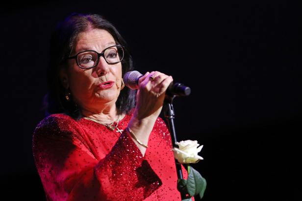 Eine ältere Frau mit Brille singt mit einem Mikrofon und hält eine weiße Rose.