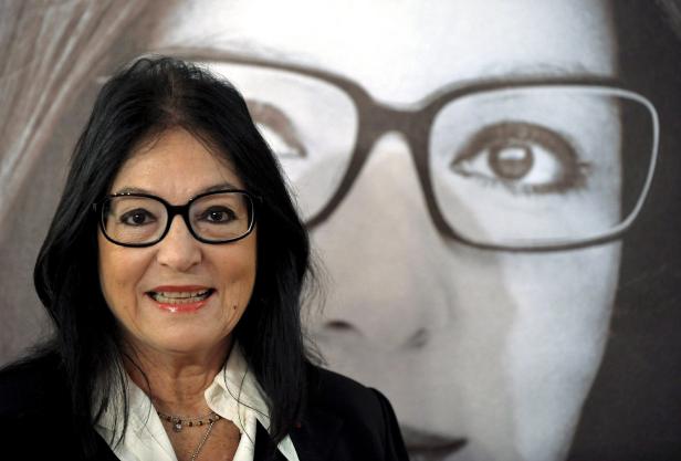Eine Frau mit Brille lächelt vor einem Porträt mit einer Person, die ebenfalls eine Brille trägt.