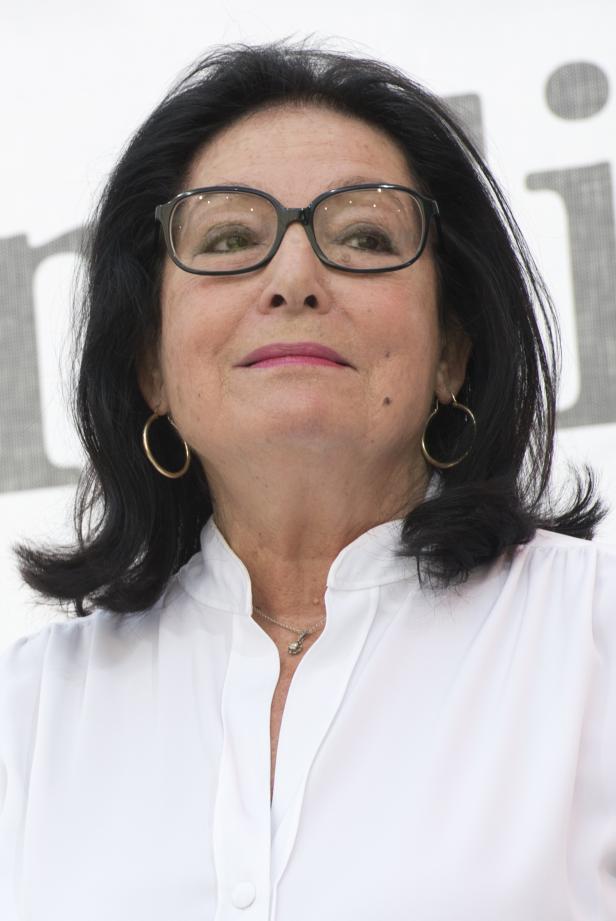 Eine Frau mit dunklen Haaren, Brille und weißer Bluse lächelt.
