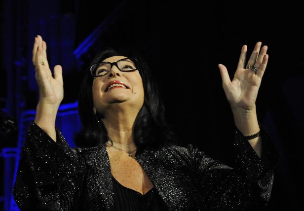 Eine Frau mit Brille und glitzernder Jacke hebt lächelnd die Hände.