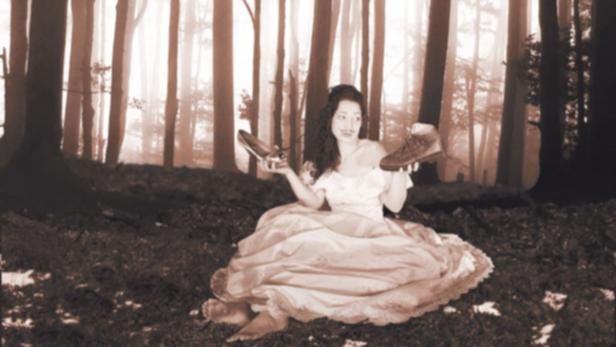 Eine Frau im Kleid sitzt barfuß im Wald und hält zwei verschiedene Schuhe hoch.
