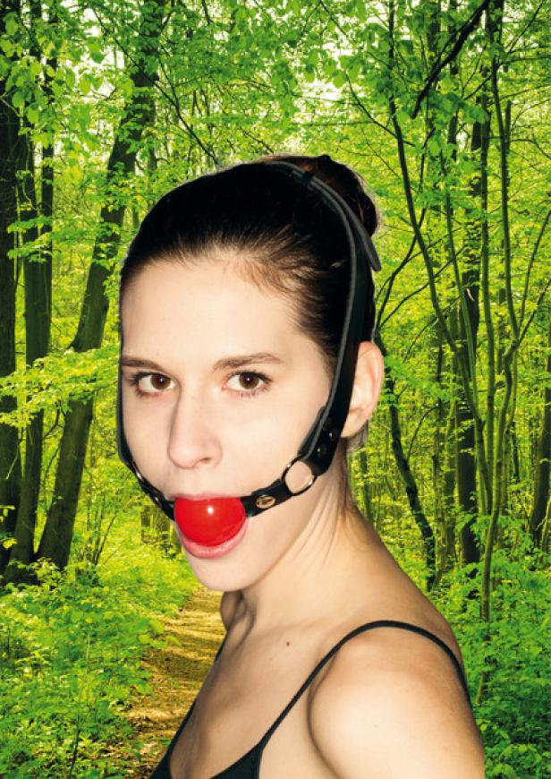 Eine Frau mit einem Knebel im Mund steht vor einem grünen Wald.