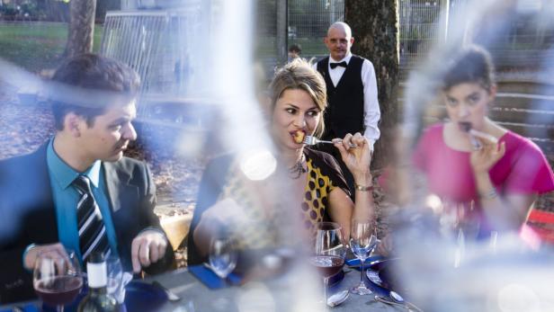 Drei Personen sitzen an einem Tisch im Freien und essen, während ein Kellner im Hintergrund steht.