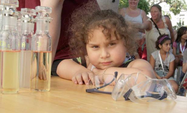 Ein kleines Mädchen lehnt an einem Tisch mit Schutzbrillen und Flaschen mit Flüssigkeiten.
