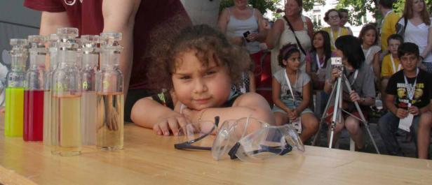 Ein Mädchen liegt auf einem Tisch mit bunten Flüssigkeiten in Flaschen und einer Schutzbrille.