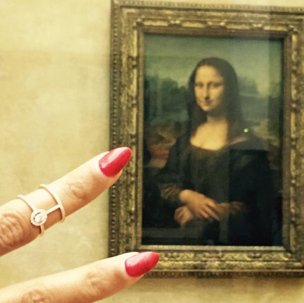 Ein Finger mit rotem Nagellack und Ring vor dem Gemälde der Mona Lisa.