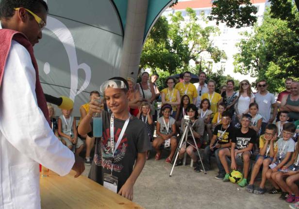Ein Junge mit Schutzbrille hält eine Flasche mit blauer Flüssigkeit in der Hand, umgeben von Zuschauern.