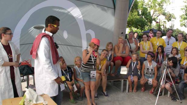 Ein Mann im Laborkittel demonstriert ein Experiment vor einer Gruppe von Kindern und Erwachsenen.