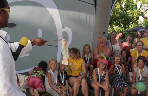 Ein Mann im weißen Kittel präsentiert mit einer Zange ein Tuch vor einer Gruppe von Kindern und Erwachsenen.