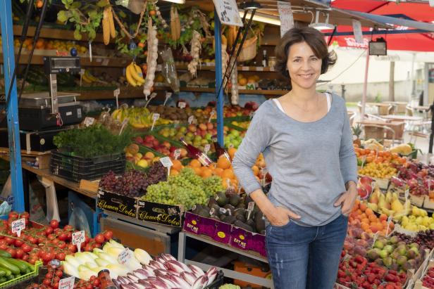 Eine Frau steht lächelnd vor einem Marktstand voller Obst und Gemüse.