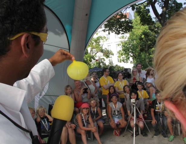 Ein Mann im weißen Kittel zeigt einen gelben Ballon vor einer Gruppe von Kindern.