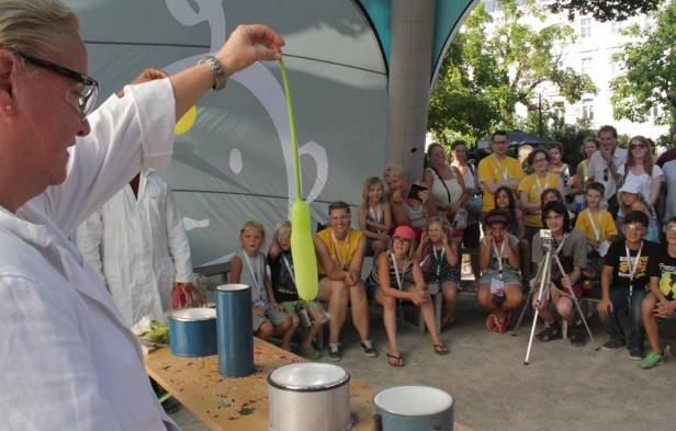 Eine Frau im Laborkittel führt vor einer Gruppe von Kindern ein Experiment mit einem Ballon durch.