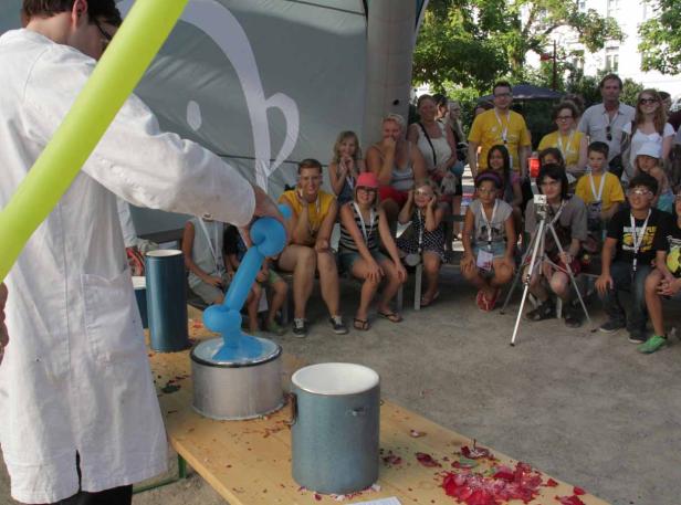 Ein Mann im Laborkittel formt einen blauen Ballon vor einer Gruppe von Kindern.