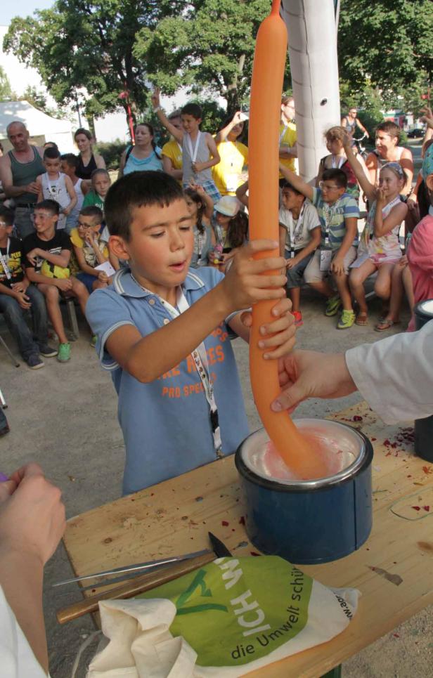 Ein Junge taucht einen langen, orangefarbenen Ballon in einen Behälter mit einer weißen Substanz.