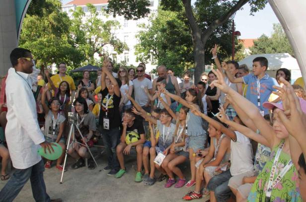 Eine Gruppe Kinder sitzt draußen und hebt die Hände, während ein Mann mit Brille und Laborkittel daneben steht.