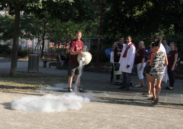 Ein Mann gießt eine rauchende Flüssigkeit aus einem Gefäß, während eine Gruppe von Menschen zusieht.