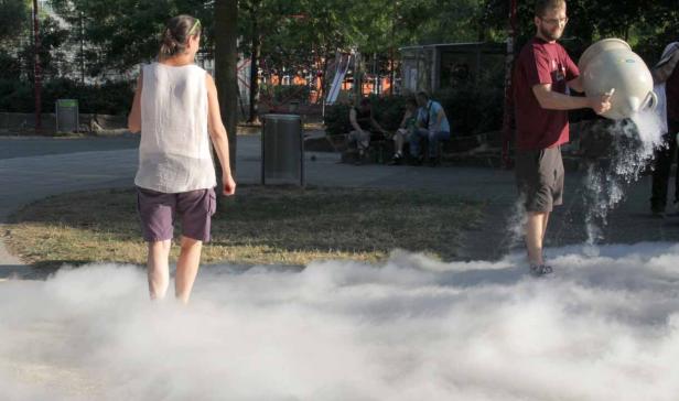 Ein Mann gießt eine Flüssigkeit aus einem Behälter, wodurch Nebel entsteht, während eine Frau durch den Nebel geht.